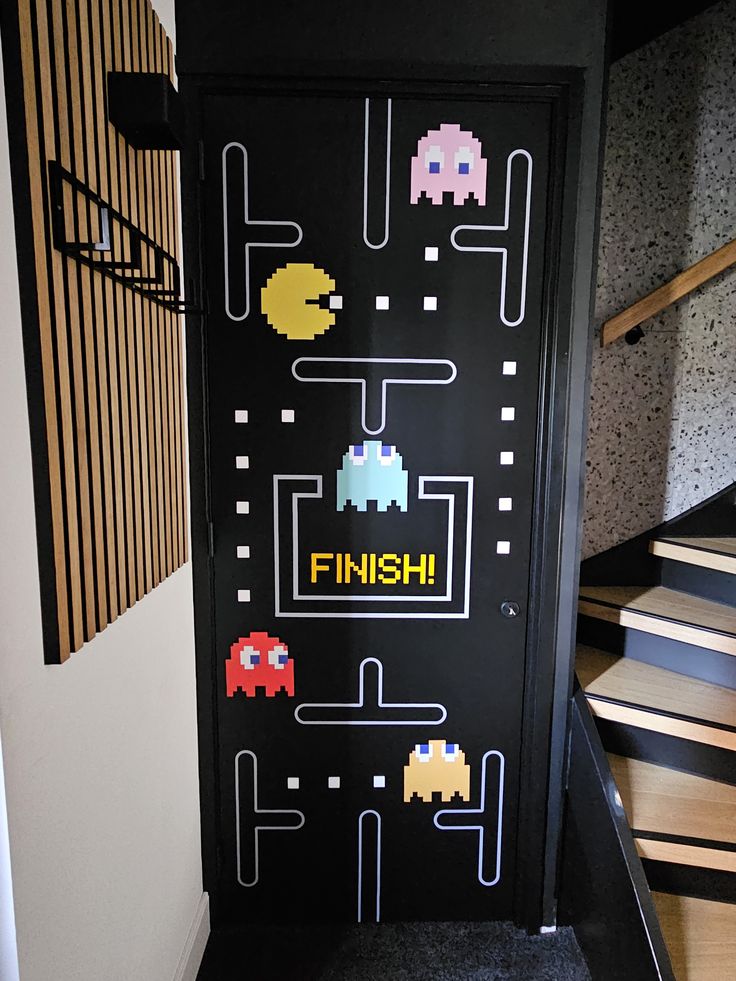 Retro Pac-Man arcade maze mural hand-painted in white and colour onto a matte black door beside a timber slat wall and staircase