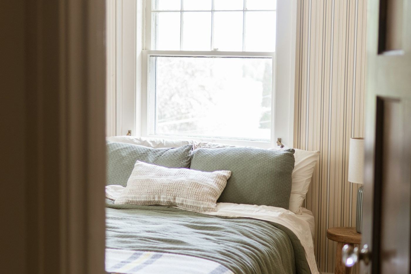 Softly striped wallpaper in a calm bedroom with sage green bedding and natural light from a window